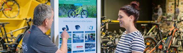 Ein eBike Fachhändler berät eine Kundin beim Kauf eines Pedelecs vor einer Infotafel von Bosch eBike Systems